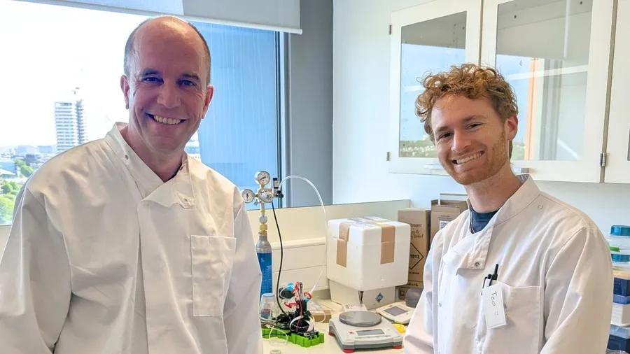 Gerrit Niezen and Teo smiling in white lab coats in an Imperial lab, with a small bioreactor set-up and a CO₂ cylinder on the bench between them